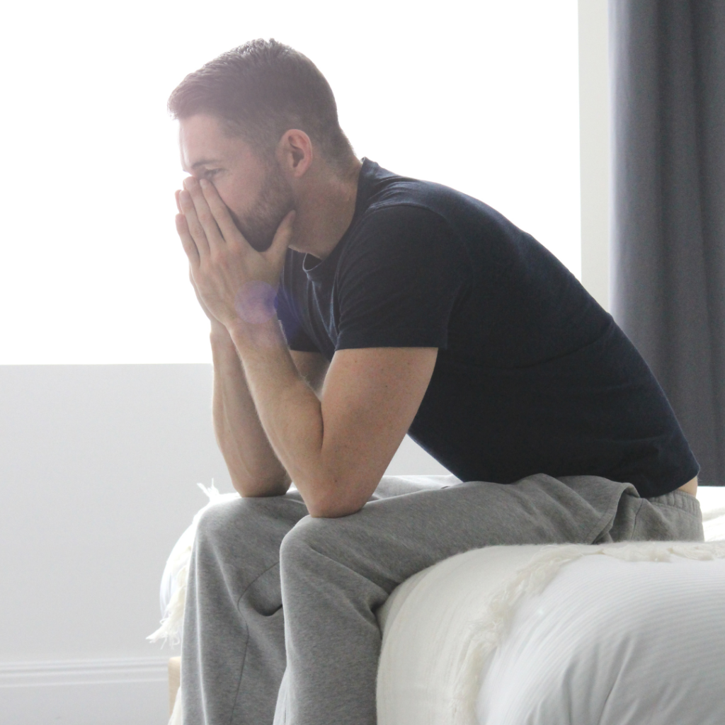 A man with OCD and Substance Use Disorder looks dejected as he sits at the edge of the bed, his face in his hands.